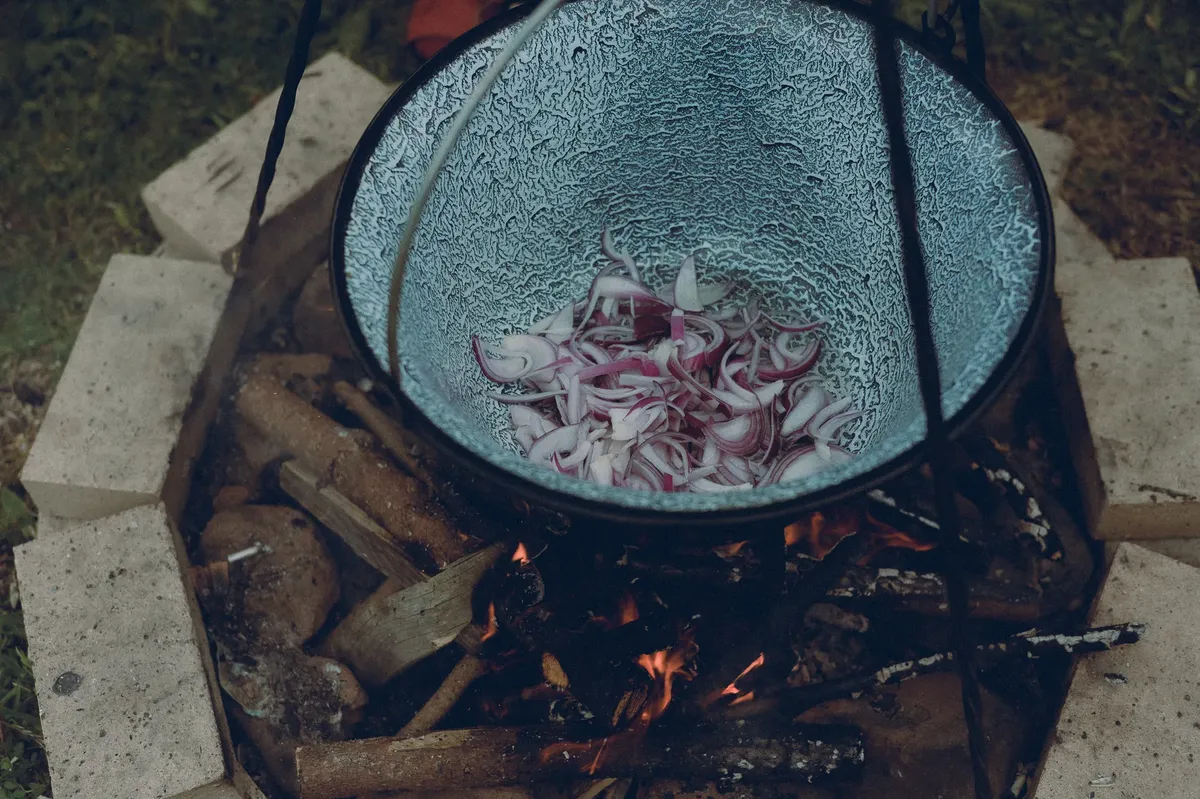 Sliced red onions cooking over an open fire in a cast iron pot outdoors.